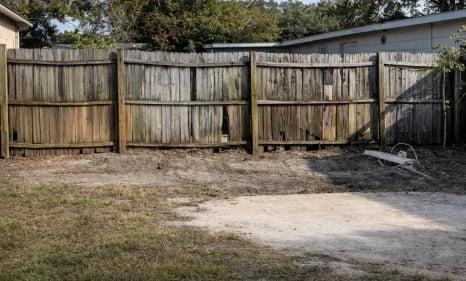 Weathered wooden fencing with multiple gate sections surrounding a gravel driveway near a house with trees in background