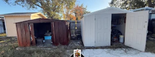 Shed demolition project - two storage sheds in a yard, one brown-red on the left with open door and one white on the right with closed doors, surrounded by grass and trees