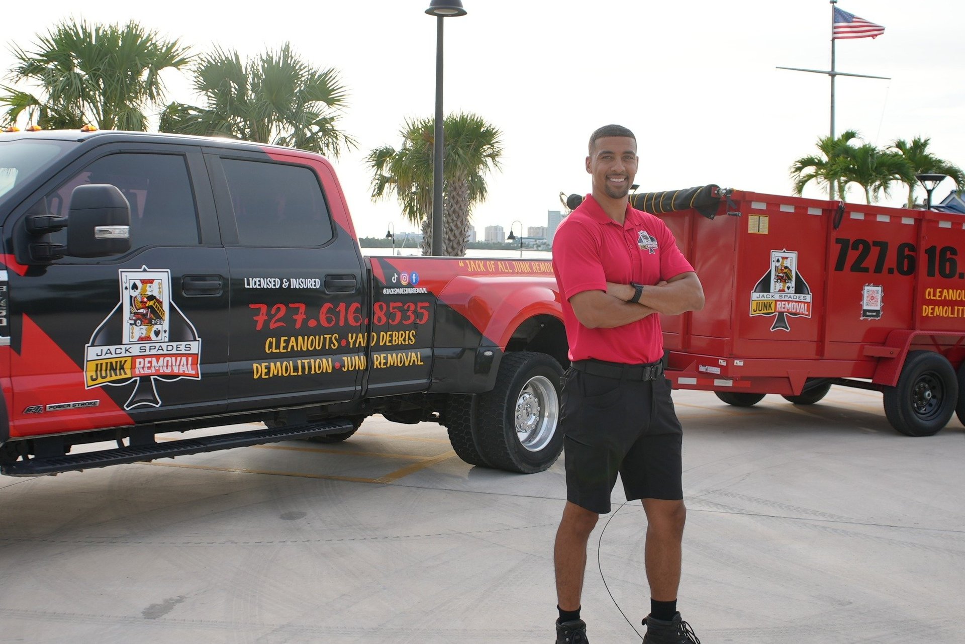 Man in red polo shirt standing in front of red service vehicle and black truck in parking lot with palm trees